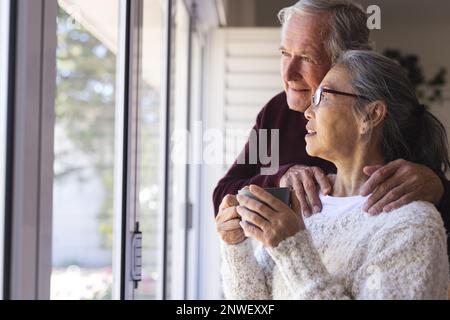 Happy senior diverse couple embracing and looking through window. Spending quality time, lifestyle, domestic life and retirement concept. Stock Photo