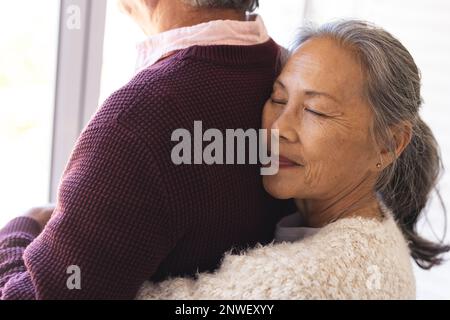 Happy senior diverse couple embracing and looking through window. Spending quality time, lifestyle, domestic life and retirement concept. Stock Photo