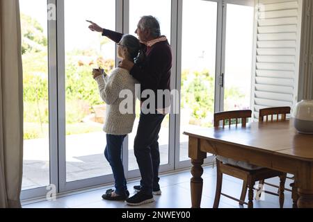 Happy senior diverse couple embracing and looking through window. Spending quality time, lifestyle, domestic life and retirement concept. Stock Photo