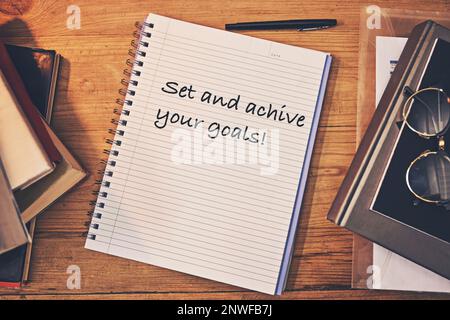 Inspiration goes a long way to being productive. Shot of a creative workstation with a notebook that has the words set and achieve you goalswritten on Stock Photo