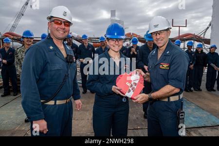 SAN DIEGO (Feb. 14, 2023) Command Master Chief Jason Ortega, left, and Capt. Aaron Taylor, right, command master chief and commanding officer of amphibious assault ship USS Essex (LHD 2), present Seaman Naomi Johnson with a prize for earning first place in a Valentine’s Day boot shining competition aboard Essex, Feb. 14, 2023. Essex is dry docked in San Diego conducting a maintenance period to upgrade and refurbish many key systems aboard. Stock Photo