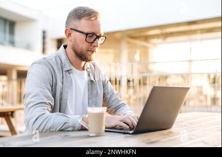 Focused smart caucasian man with a glasses and in a casual stylish clothes, IT specialist, developer, programmer, working in a laptop outdoors, developing a new program, writing code Stock Photo