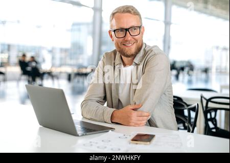 Photo of a successful caucasian man with glasses, in stylish casual wear, IT specialist, company seo, programmer, digital developer, sits at a desk with a laptop in modern office, looking away, smiles Stock Photo