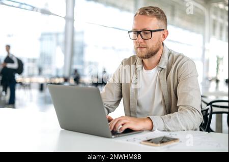 Concentrated busy caucasian man with glasses, in stylish casual wear, IT specialist, software tester, programmer, digital developer, works in a laptop in modern office, testing a new program Stock Photo