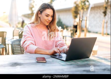 Positive confident successful gorgeous caucasian young woman, seo, product manager, working online in a laptop while sitting at a table outdoors, exploring the potential of a new digital project,risks Stock Photo