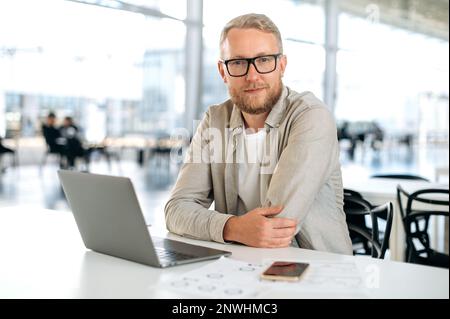 Clever successful caucasian man, with glasses, in stylish casual wear, IT specialist, company seo, programmer, digital developer, sits at a desk with a laptop in coworking, looks at camera, smiles Stock Photo