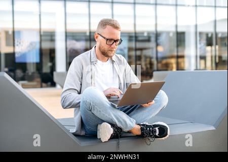 Clever focused busy caucasian man with a glasses, in a casual clothes, IT specialist, developer, programmer, sit with a laptop outdoors, developing a new program, writing code, working online Stock Photo