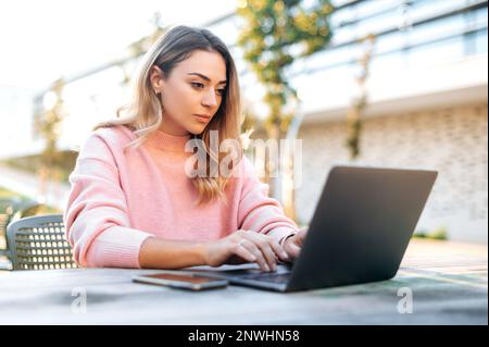 Focused smart confident successful lovely caucasian young woman, seo, freelancer, company worker, working in a laptop sitting at a table outdoors, exploring the potential of a new digital project Stock Photo