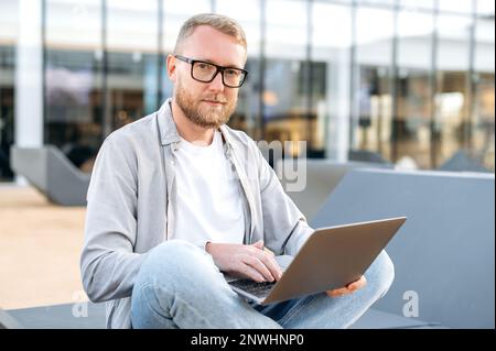 Positive attractive clever caucasian man with a glasses, in a casual clothes, IT specialist, developer, programmer, sit with a laptop outdoors, developing a new program, writing code, smiles at camera Stock Photo