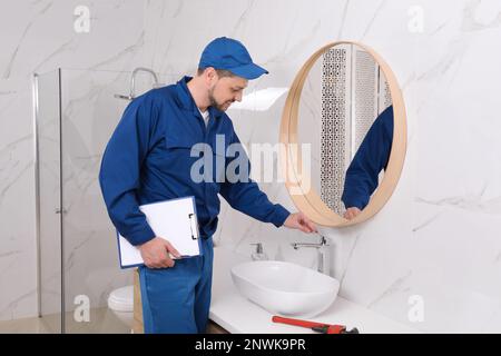Professional plumber with clipboard checking water tap in bathroom Stock Photo