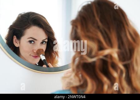 young woman powdering her nose with make-up brush seen in bathroom mirror, selective focus Stock Photo