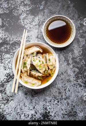 Asian dumplings in broth, bowl, chopsticks, soy sauce, rustic stone background. Top view. Chinese dumplings for dinner. Closeup. Traditional Stock Photo