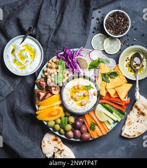 Meze platter with hummus, yoghurt dip, assorted snacks. Hummus in bowl, vegetables sticks, chickpeas, olives, pita, chips. Plate with Middle Stock Photo