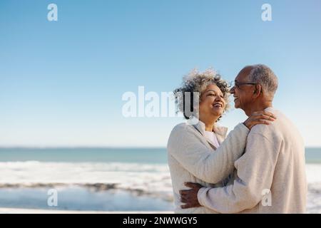 Romantic senior couple smiling happily while standing together on a wooden foot bridge at the beach. Cheerful elderly couple enjoying a refreshing sea Stock Photo