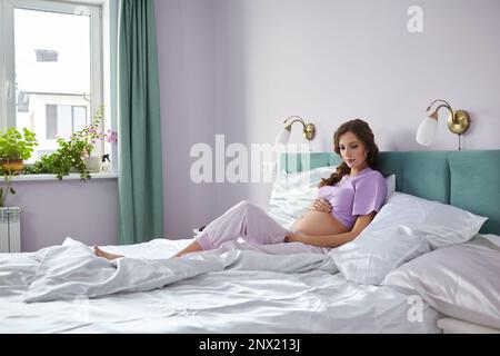 A pregnant woman sits in bed of her domestic bedroom. Female dressed in pajama enjoying her pregnancy state. Stock Photo