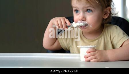 Young Kid Eating Blend Mashed Feed Sitting in High Chair. Baby Weaning. Little Girl Learning to Eat Yogurt, Feeding Himself. Small Hand with Spoon. Br Stock Photo