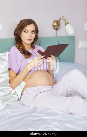 A pregnant woman lies in bed of her domestic bedroom. Female dressed in pajama reads an e-book. Stock Photo