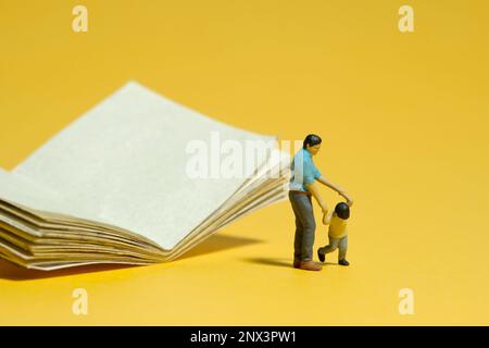 Miniature people toy figure photography. Kids refuse to read a book concept. A father and son standing in front of opened book. Isolated yellow backgr Stock Photo