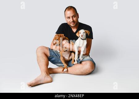 Handsome man dressed in black t-shirt with his little dogs in his arms poses on gray background. Stock Photo