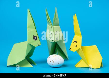 Three paper origami bunnies and an Easter egg on a blue background. Crafts with your own hands for Easter Stock Photo