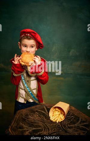 Happy cheerful little boy wearing costume of medieval page boy and prince eating hamburger with french fries over dark green background. Stock Photo