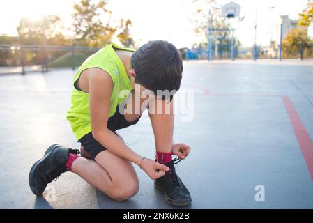 Close-up of a boy tying his shoelaces on an urban basketball court at sunset. Stock Photo