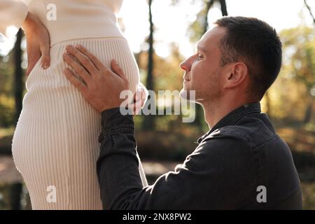Cropped shot of happy young husband enjoying strokes, talks and kisses tummy of wife, waiting for birth baby outdoors in sunbeams in the city park. Ma Stock Photo