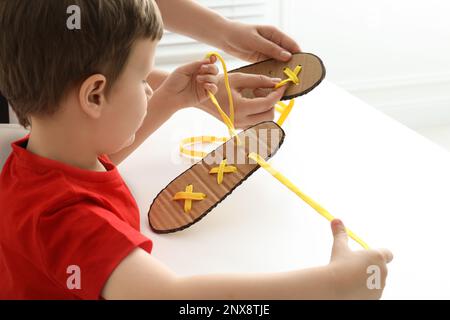 Mother teaching son to tie shoe laces using training cardboard template at white table, closeup Stock Photo