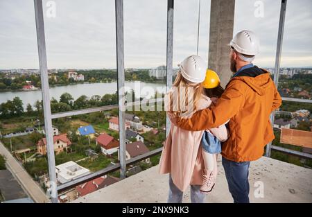 Back view of parents with daughter standing inside apartment building under construction. Man and woman with child wearing building helmets while enjoying city view from future home. Stock Photo