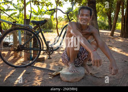 A middle aged man squats on a large stone next to his bicycle at a village near Siem Reap in Cambodia. Stock Photo