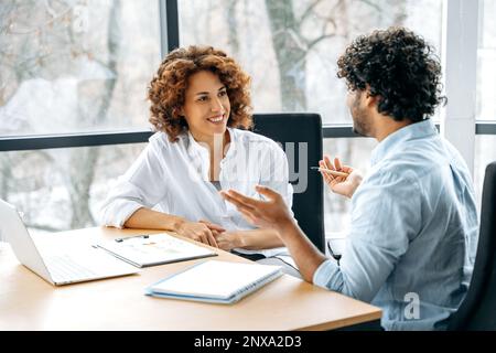 Two positive confident motivated employees of the company of different nationalities are working together on a new project in a modern office, together discussing ideas and goals, communicating Stock Photo