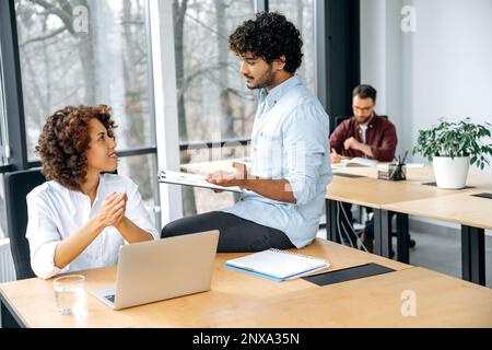 Teamwork concept. Two successful employees of the company of different nationalities are working together on a project using a laptop, in a modern office, analyzing solutions to achieve goals Stock Photo