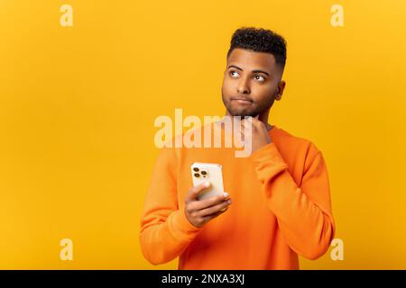 Portrait of man holding chin, standing with smartphone in hand, contemplating about software updating, choosing suitable tariffs. Indoor studio shot isolated on yellow background Stock Photo