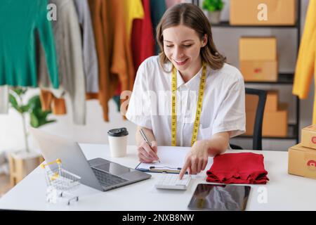 Concept of fashion and  designer,Woman fashion designer stylish with measuring tape on neck sitting and working about fashion design using laptop whil Stock Photo