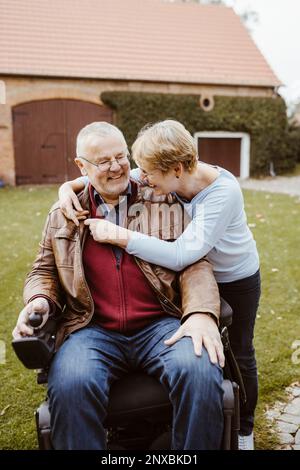 Cheerful retired senior woman hugging man with disability sitting in motorized wheelchair at back yard Stock Photo
