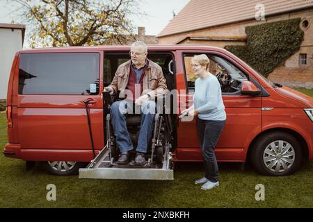 Smiling senior woman holding remote control while helping man with disability in motorized wheelchair to disembark from Stock Photo
