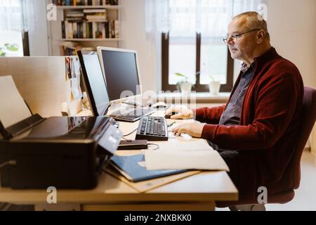 Side view of senior man typing while using computer at desk in home office Stock Photo