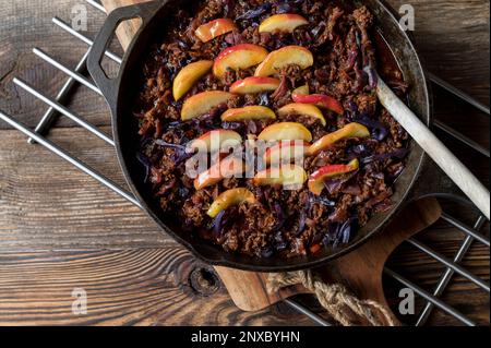 Ground beef pan with red cabbage and cooked apple topping in a cast iron pan isolated on wooden table. Top view with copy space Stock Photo