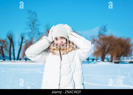 Cute woman squints from sun, touching white cap on her head with hands. Winter sunny day. Stock Photo