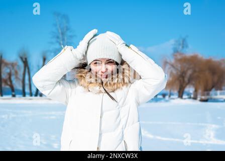 Cute woman squints from sun, touching white cap on her head with hands. Winter sunny day. Stock Photo