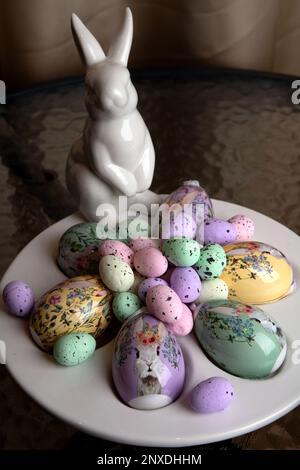 photo large and small eggs lying on a plate and next to a white rabbit on a glass table Stock Photo