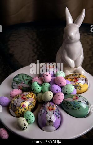 photo bright decorative large and small eggs lie on a special plate and next to a white rabbit on a round glass table Stock Photo