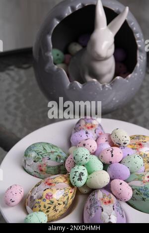 photo many different colored decorative eggs of different sizes lying on a white plate and a white rabbit standing on a round glass table Stock Photo