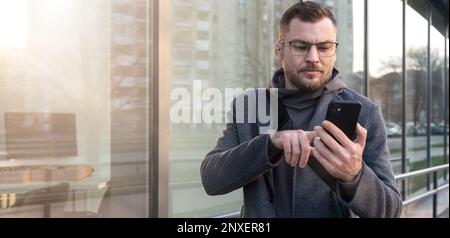 Man entrepreneur wearing glasses using his smartphone outdoors in city, man reading email massage on mobile phone. Stock Photo
