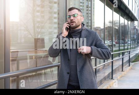 A young businessman in glasses stands near the business center and talks on a mobile phone. Stock Photo