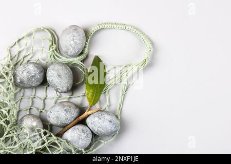 Grey Easter eggs with a sprig and a magnolia leaf in a green string bag on a grey background. Minimalism, copy space, flat lay Stock Photo