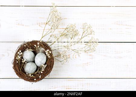 A brown nest of twigs with three gray Easter eggs, feathers and sprigs of gypsophila on a white wooden table. Minimalistic Easter concept Stock Photo