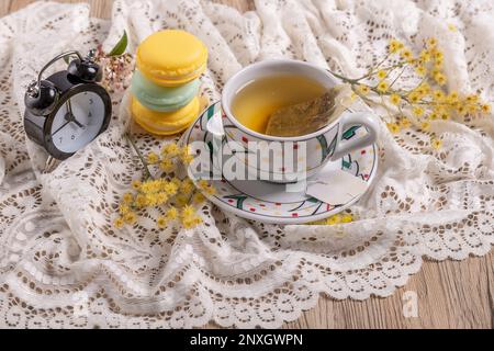 A cup of tea next to an alarm clock that strikes five o'clock on a lace tablecloth with three stacked macarons Stock Photo