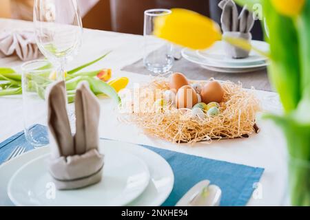 Beautiful Easter table setting with festive decor indoors. Napkin in shape of rabbit ears, colorful eggs in nest, fresh yellow tulips in vase. Sunligh Stock Photo