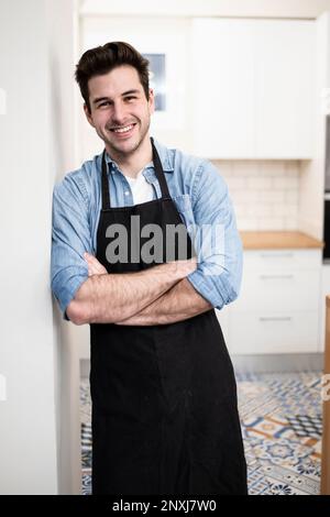 handsome young man smiling in the kitchen wearing an apron - Portrait of a cheerful confident caucasian young man looking at camera with arms crossed  Stock Photo